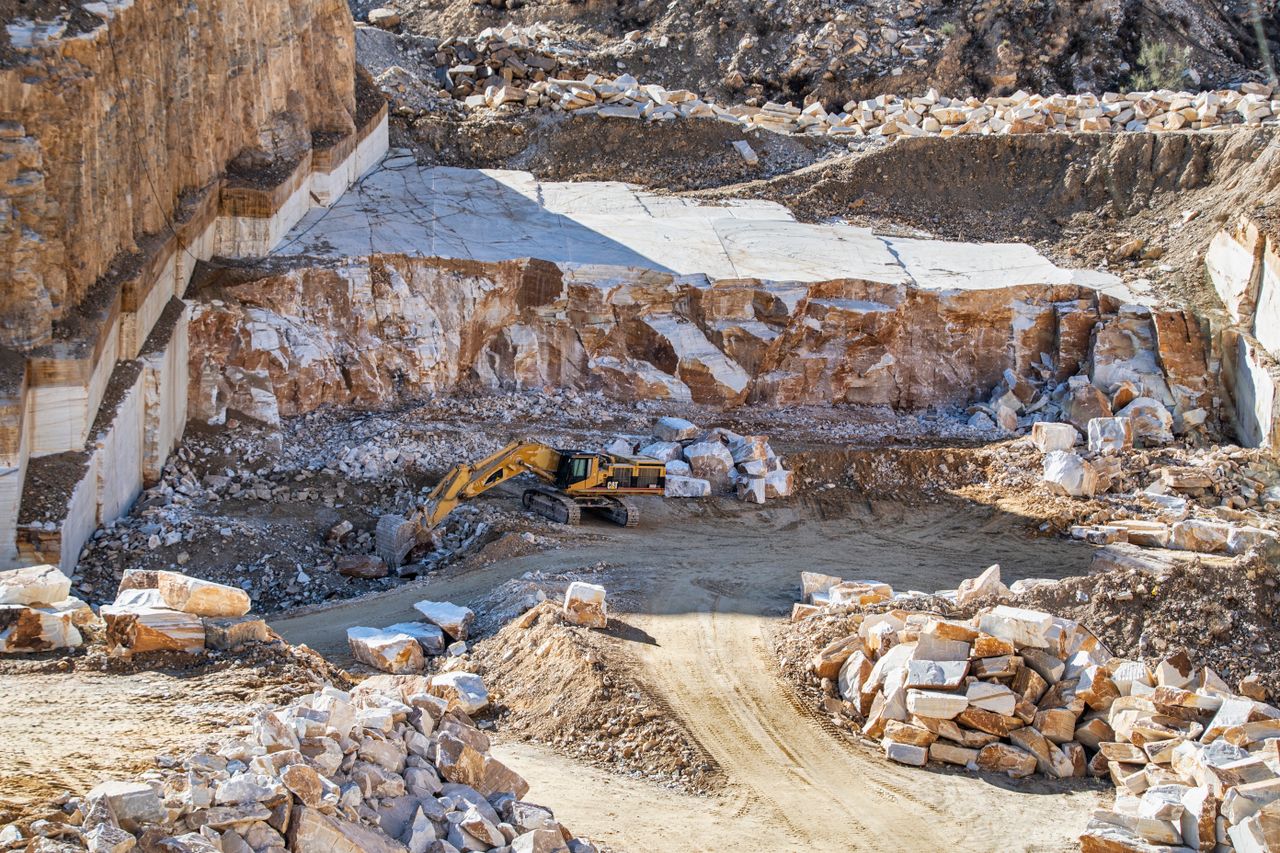 service-06 An excavator operates in a large stone quarry with rugged rocky terrain under daylight.